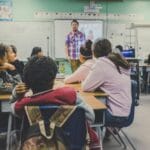 Rows of students facing a teacher talking at the front of a classroom