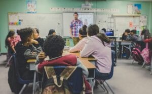 Rows of students facing a teacher talking at the front of a classroom