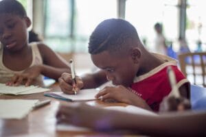 Two young children writing in notebooks with pencils at a desk